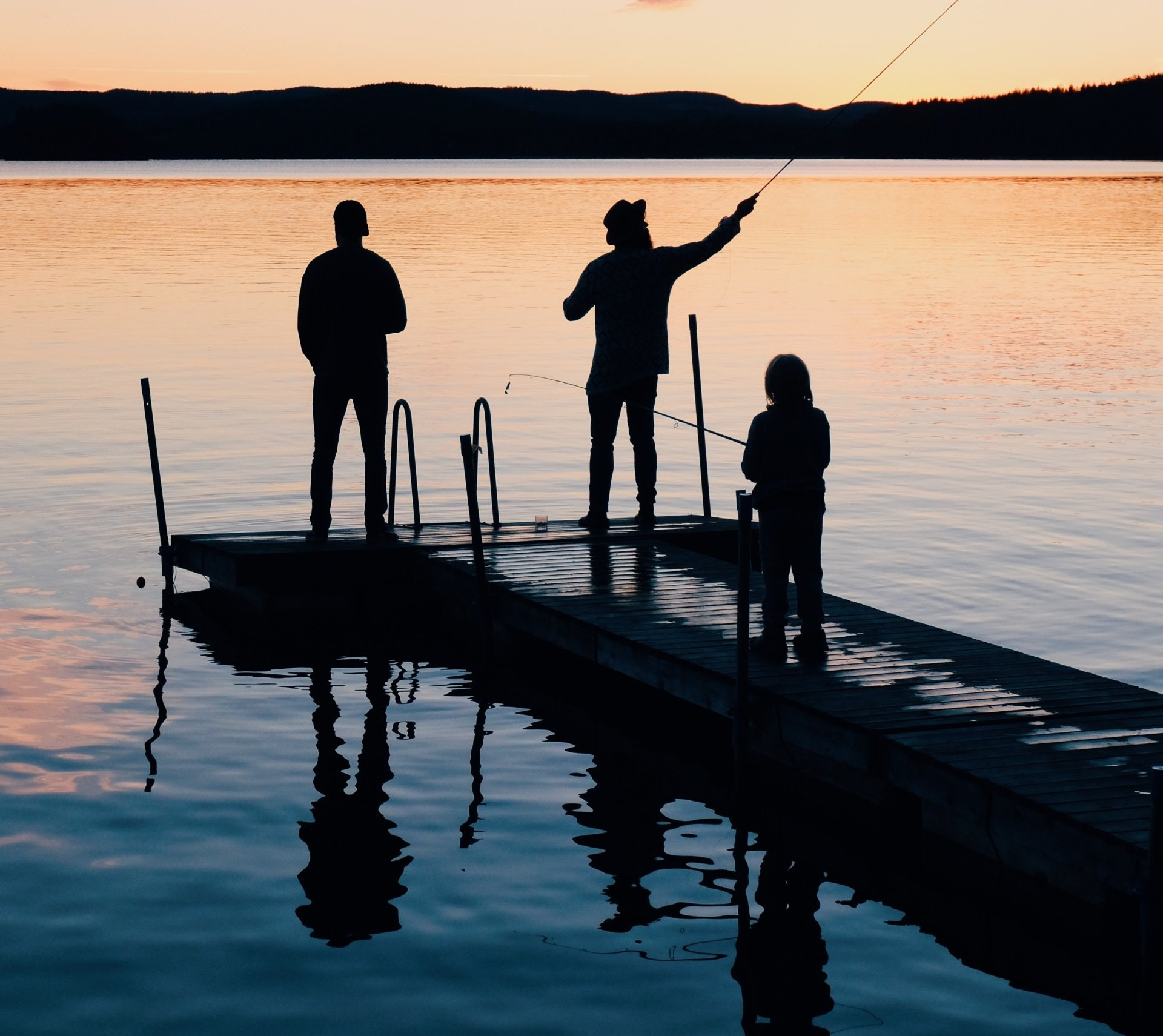 Silhouettes of three people fishing from a dock at sunset over calm water