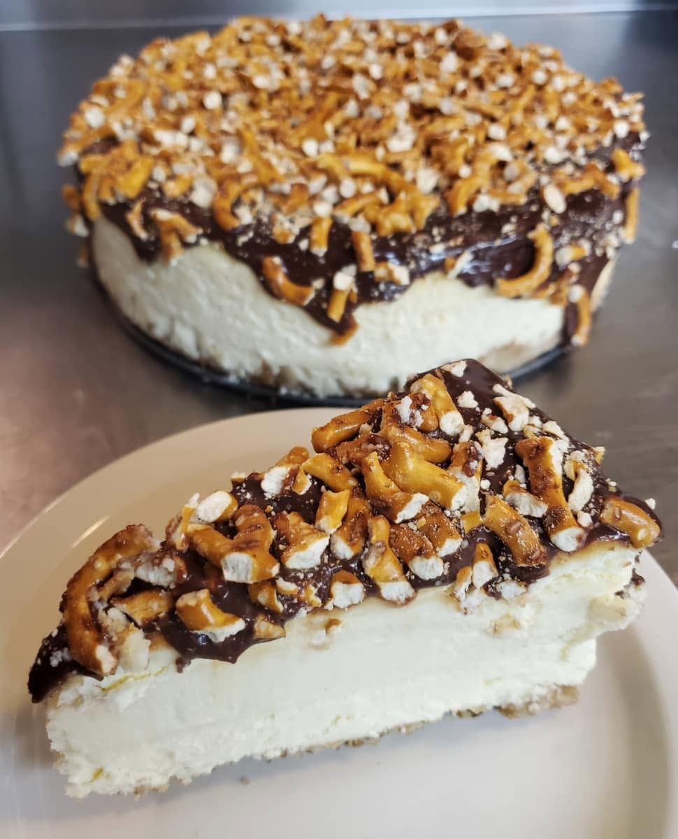 Slice of caramel nut cheesecake on a plate, with the rest of the cake in the background