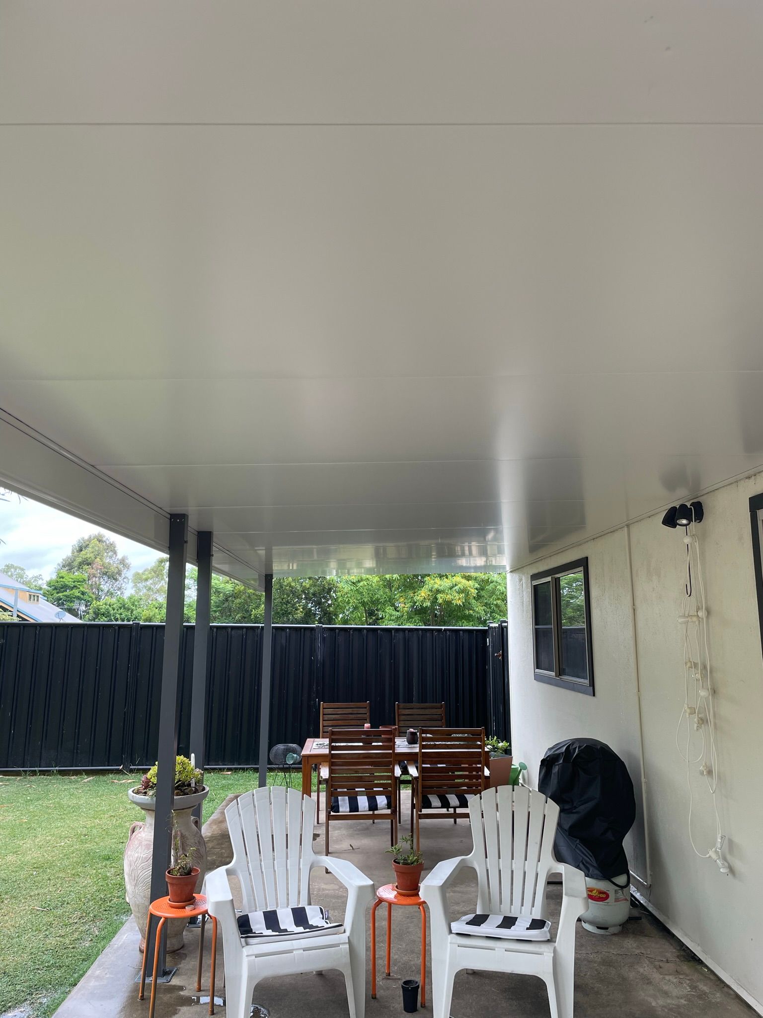 Patio With White Chairs, Wooden Table, Black Fence, and Greenery — Taylormade Constructions & Renovations In Wooroolin, QLD