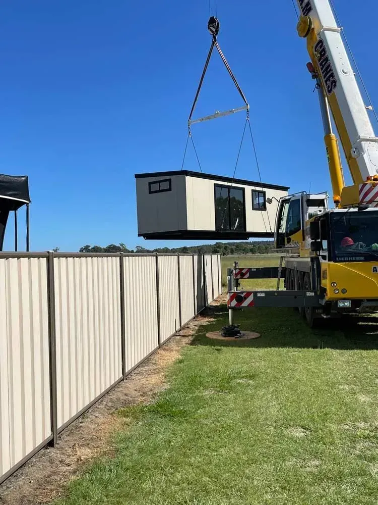 Crane Lifting a Small, Cream-colored Building Over a Beige Fence — Taylormade Constructions & Renovations In Tarong, QLD