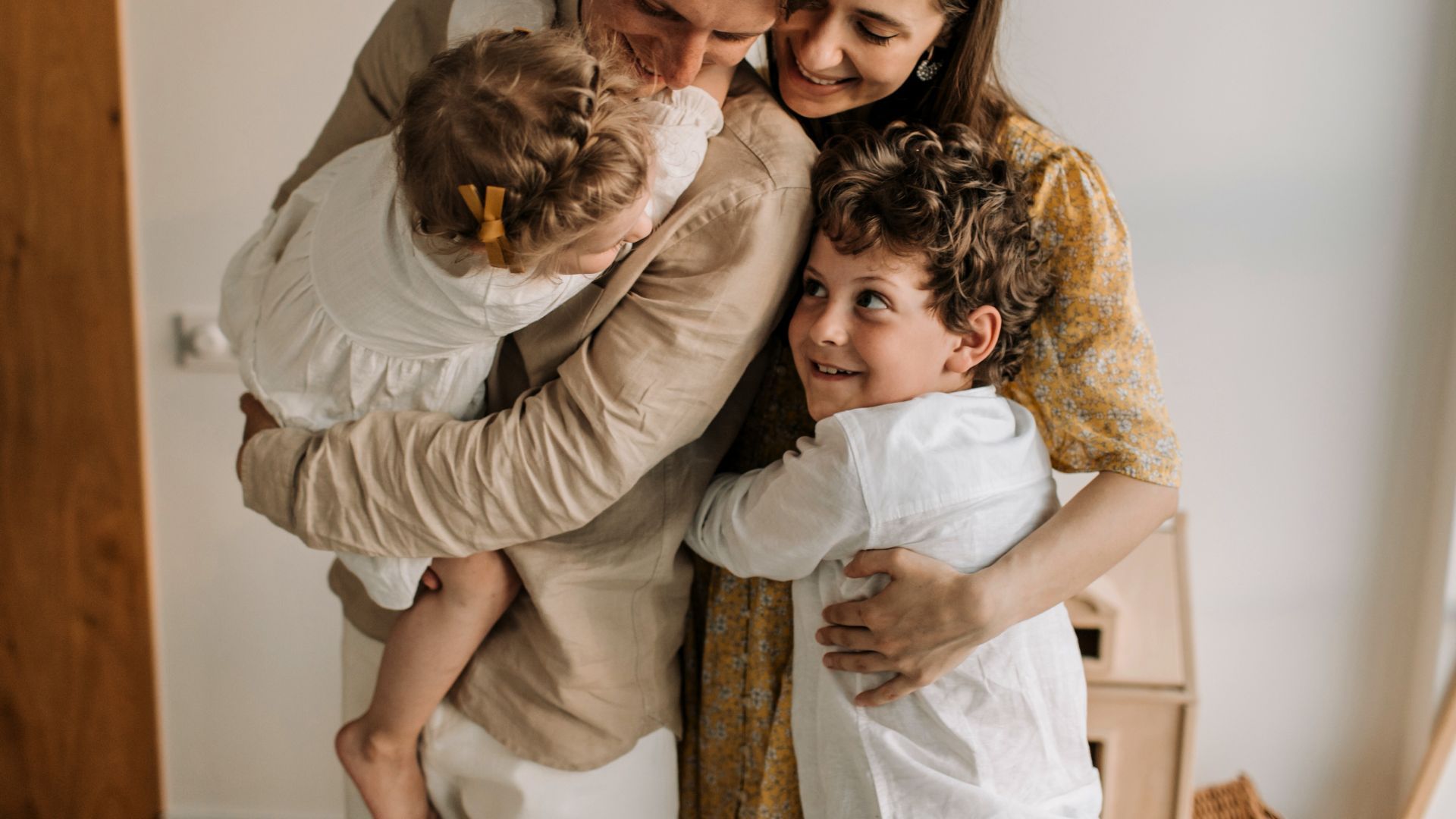 Family of four embraces: parents, a smiling boy, and a child being held, all in soft-colored clothing.