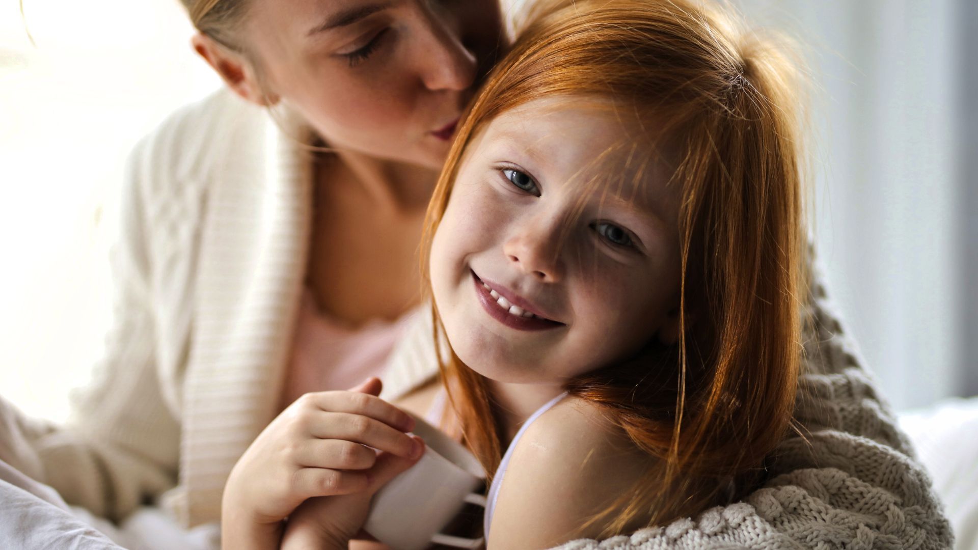 Family embraces: parents hold two children in neutral-toned room; children smile happily.