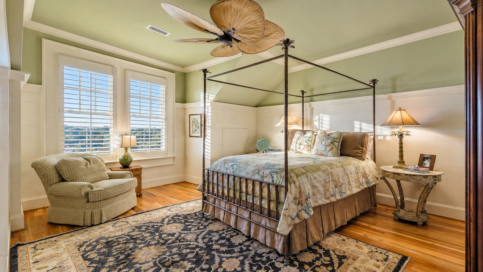 Bright bedroom with a four-poster bed, patterned rug, armchair, and ceiling fan in soft neutral tones