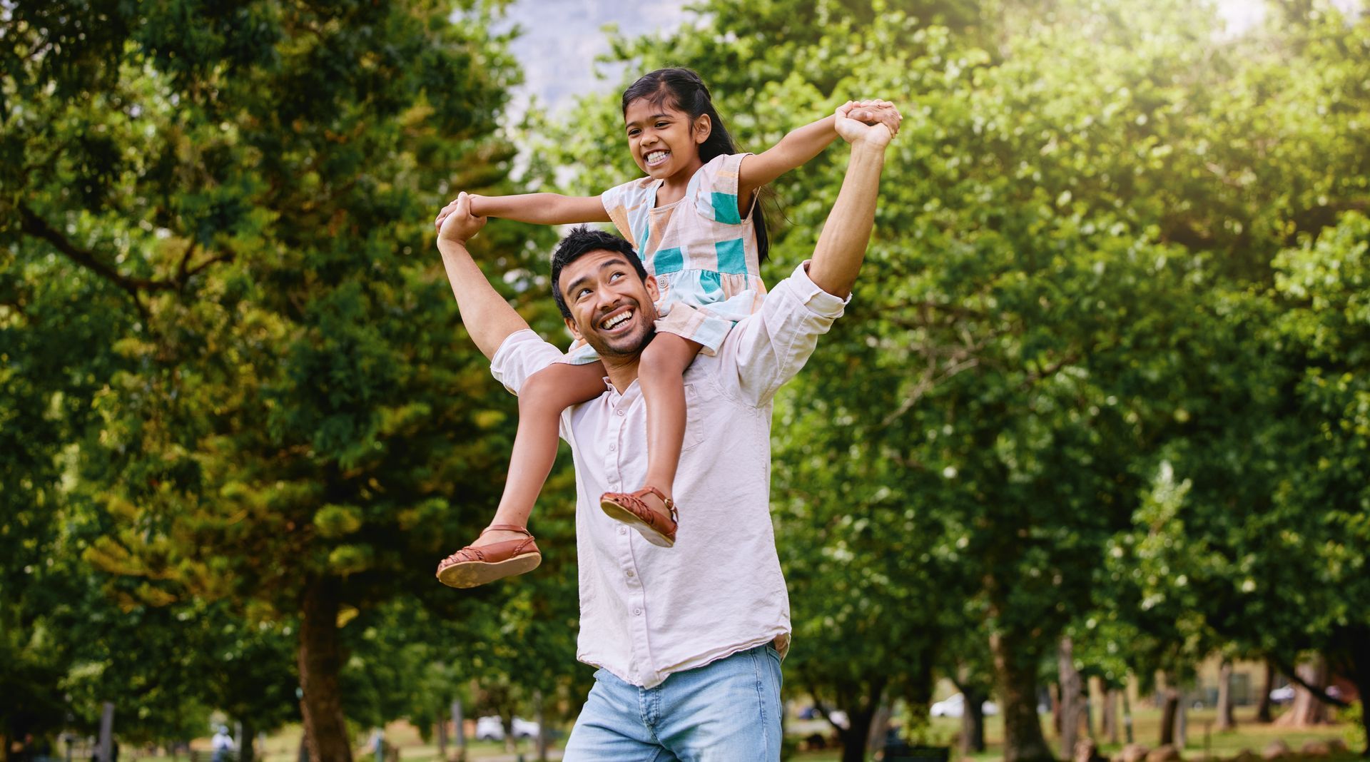 Father carries daughter on his shoulders, smiling in a park with green trees.