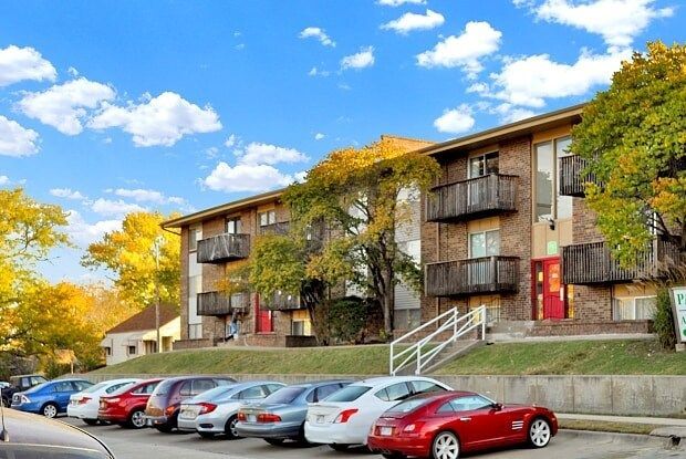 Apartment building with balconies and red doors, cars parked in front, blue sky with clouds.