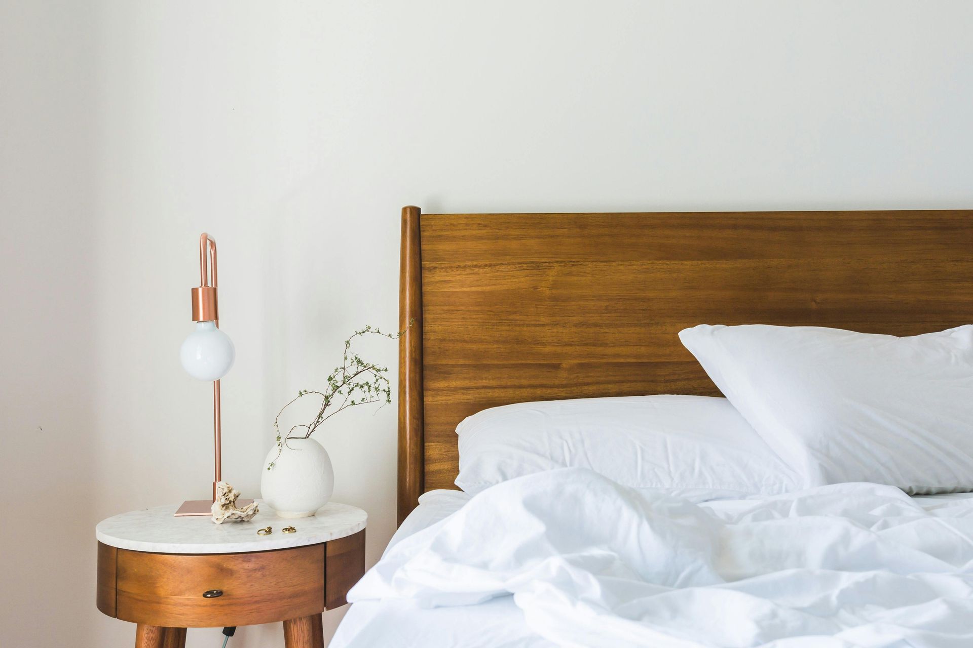 Wooden bed headboard, white bedding, nightstand with lamp and vase.