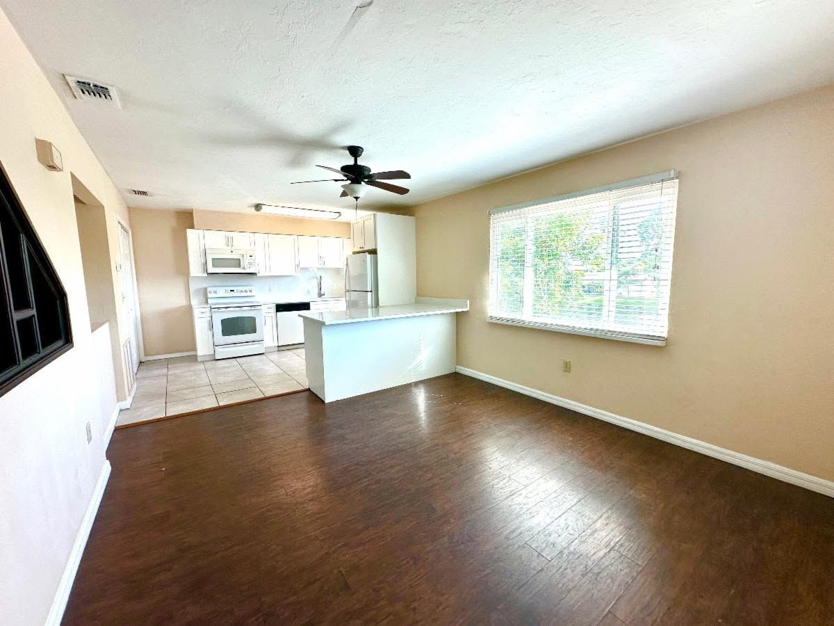 Empty living room with dark wood floors, beige walls, and open view to a white kitchen and bright window.