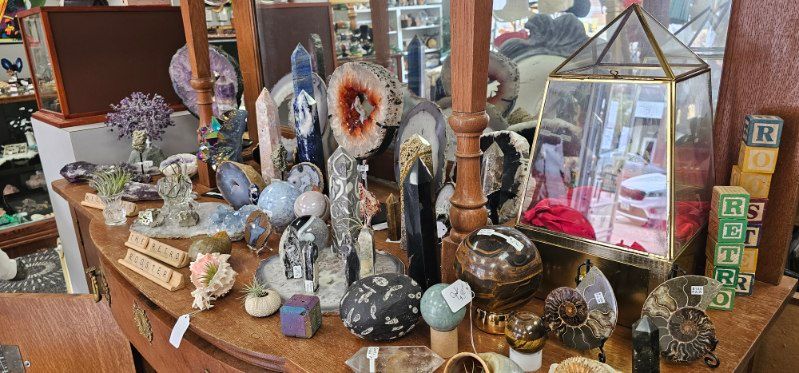 Glass and crystal décor on a wooden table in a shop, with framed art and hanging ornaments.
