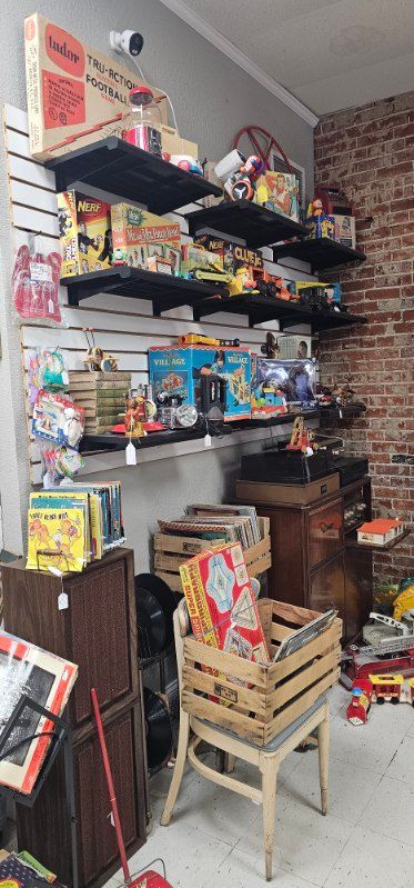 Cluttered storage room with shelves of boxes, snacks, and miscellaneous household items, plus stacked crates and a chair
