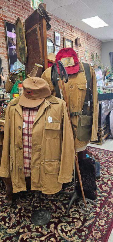 Two tan coats on mannequins in a vintage shop, one with a straw hat and plaid shirt.