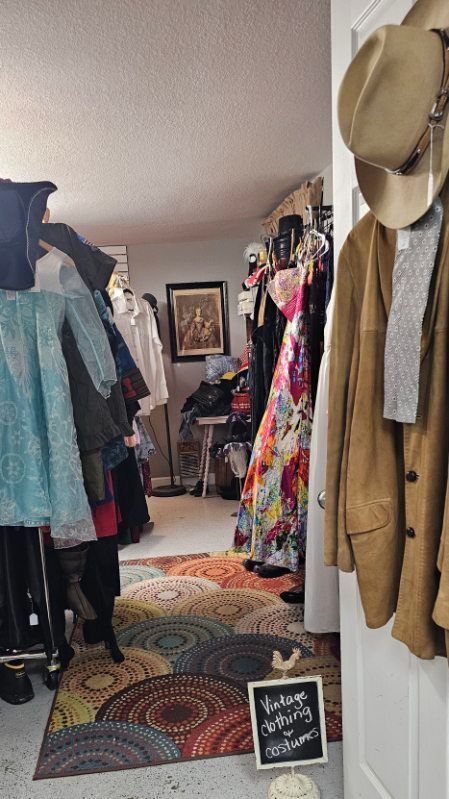Clothing racks line a narrow boutique aisle with colorful dresses, coats, and hats on the walls.