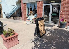 Sidewalk storefront with a sandwich board sign, potted flowers, and an outdoor stairway in sunlight.