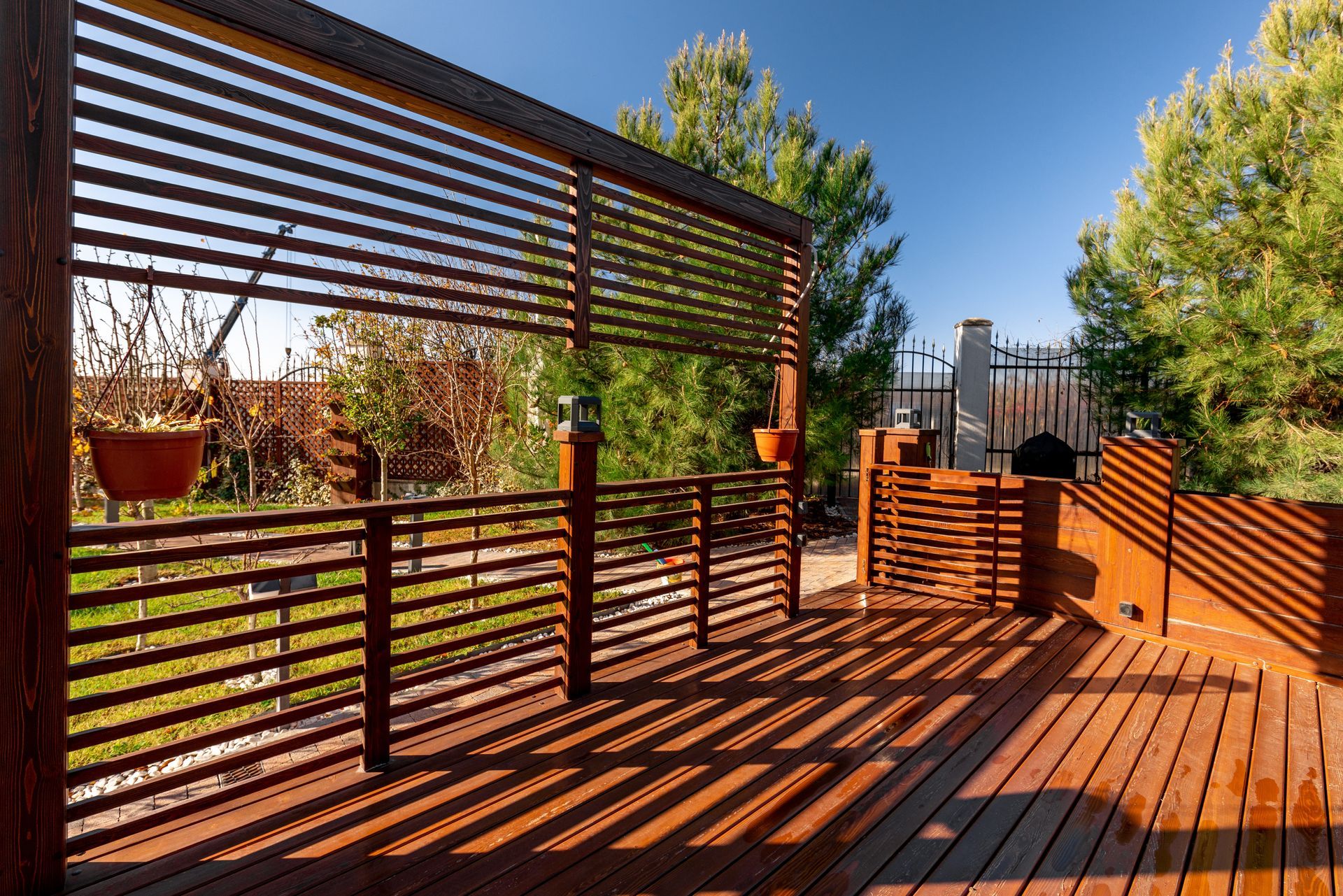 Wooden deck with slatted pergola railing, warm sunlight, potted plants, and trees beyond
