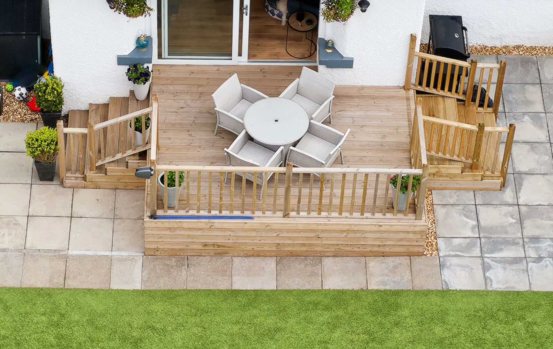 Small patio deck with a round table, white chairs, wooden fencing, and potted plants beside a lawn.