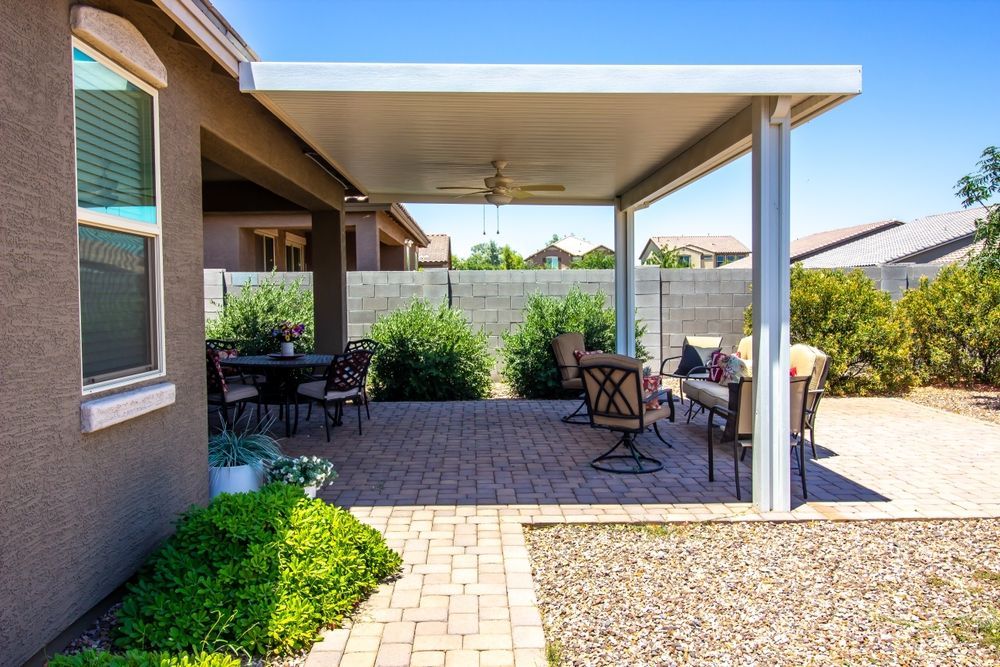 Covered patio with outdoor seating, potted plants, and a paved backyard area under bright sunlight