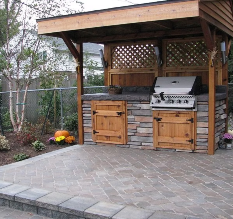 Wooden outdoor kitchen pavilion with grill, stone counter, cabinets, and paved patio in a backyard garden