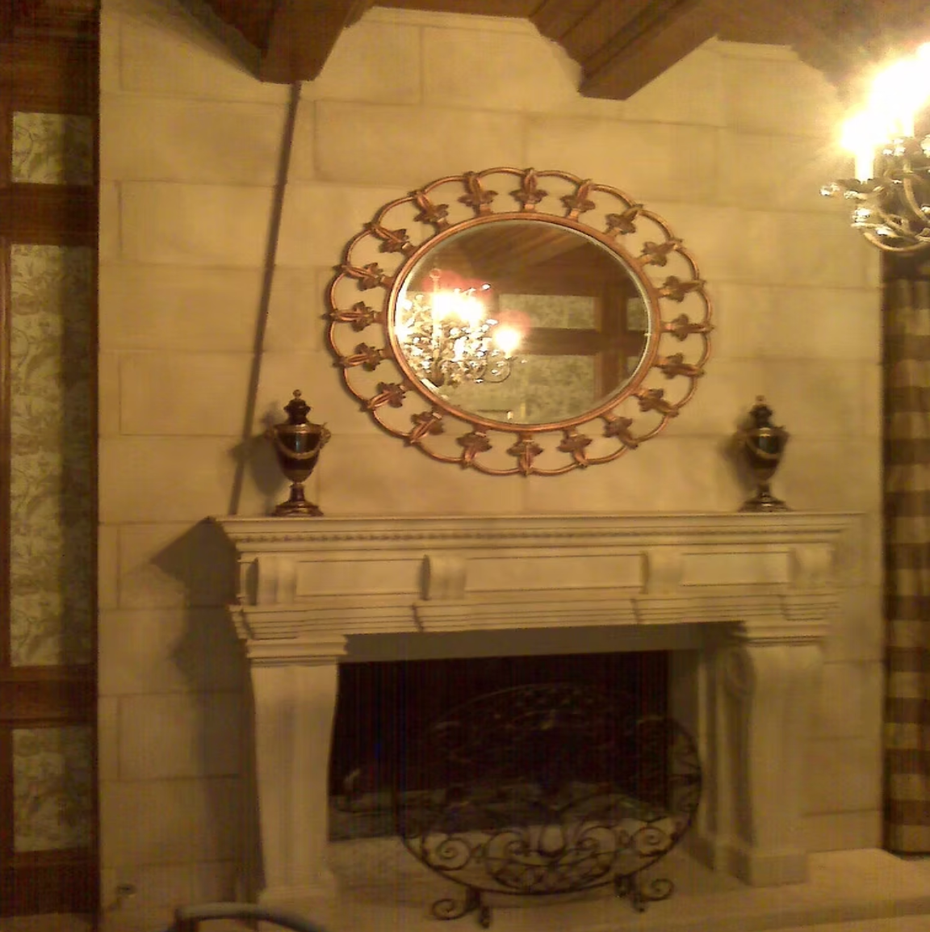 Ornate fireplace with mirror above, in a beige stone room, lit by warm chandelier light.