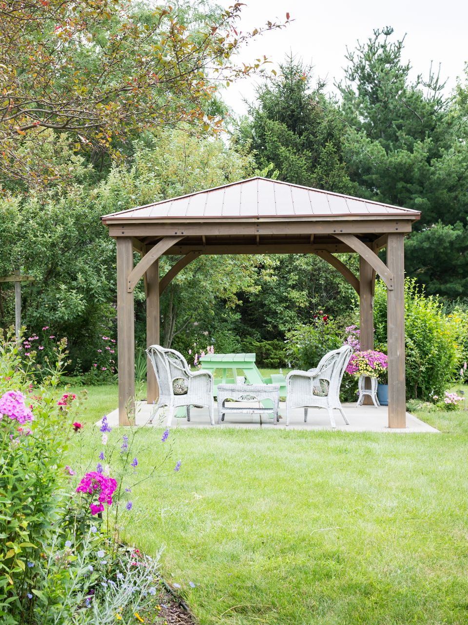 A gazebo is sitting in the middle of a lush green yard surrounded by flowers.