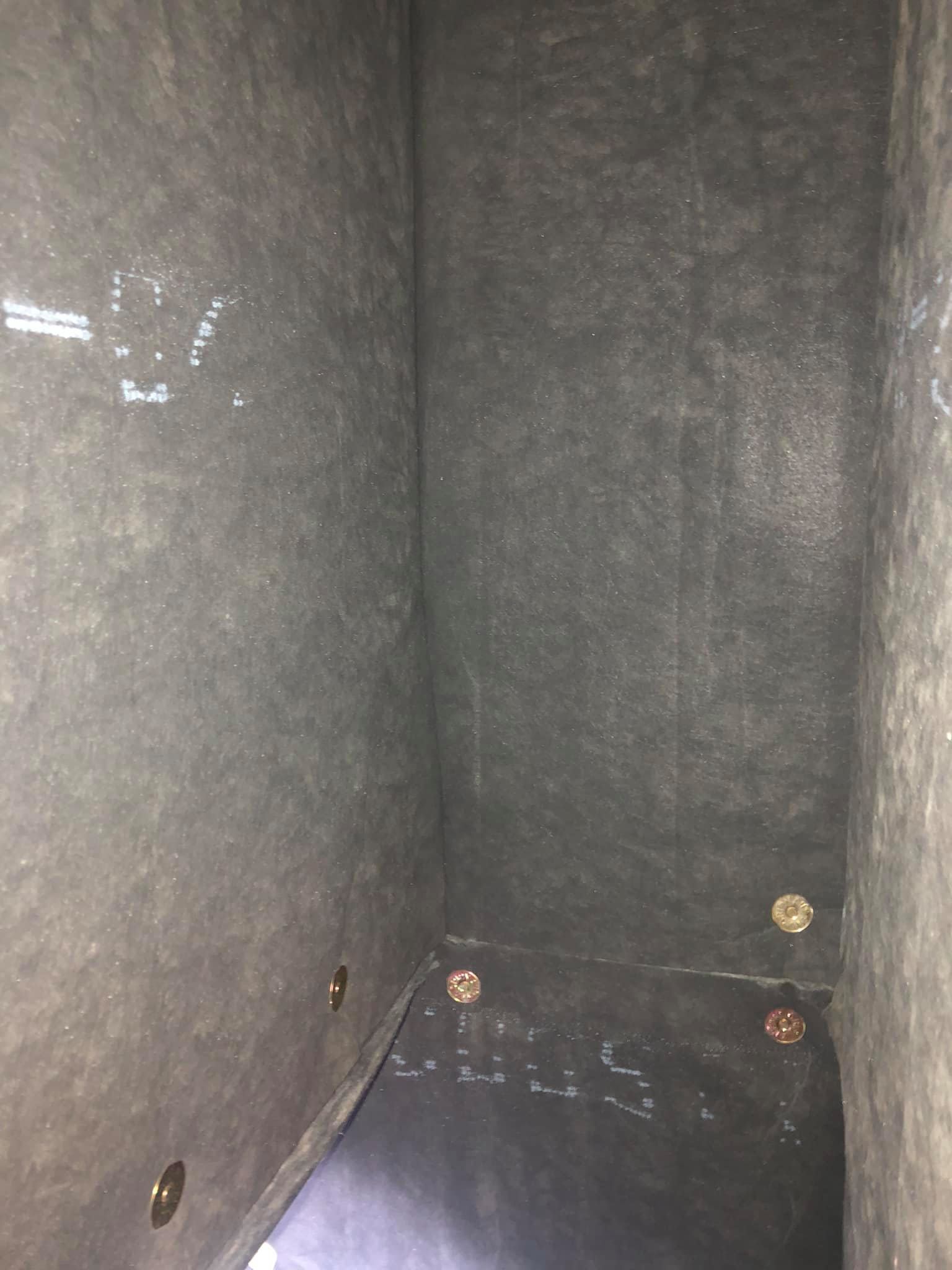 Compartiment de rangement vide, recouvert de moquette grise, avec des fixations métalliques et un sol foncé.