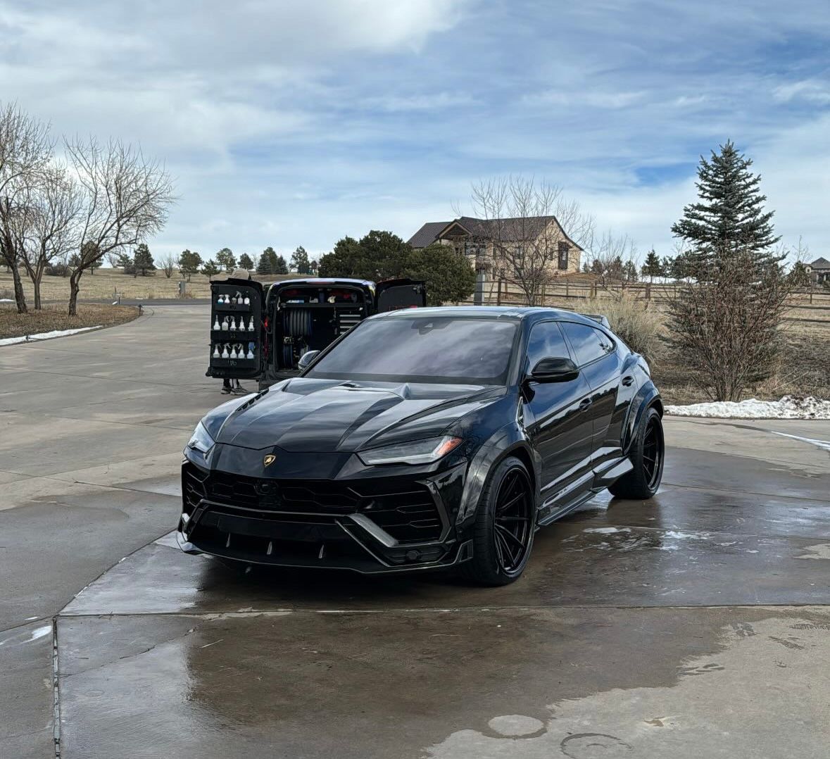 Black Lamborghini SUV parked on wet concrete with portable restrooms in background.
