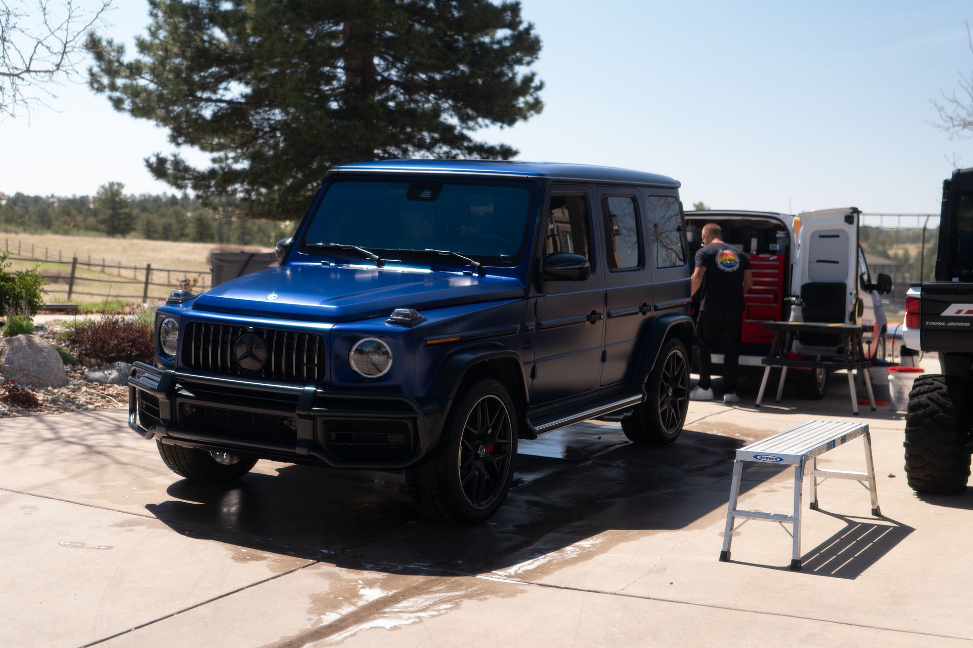 Blue SUV parked in a driveway beside a trailer, with a person standing nearby outdoors.