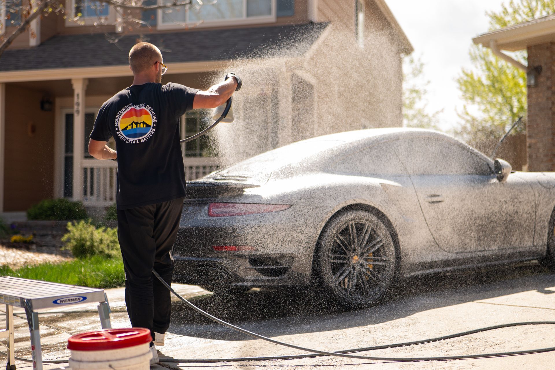Person spraying foam over a black sports car in a sunny driveway