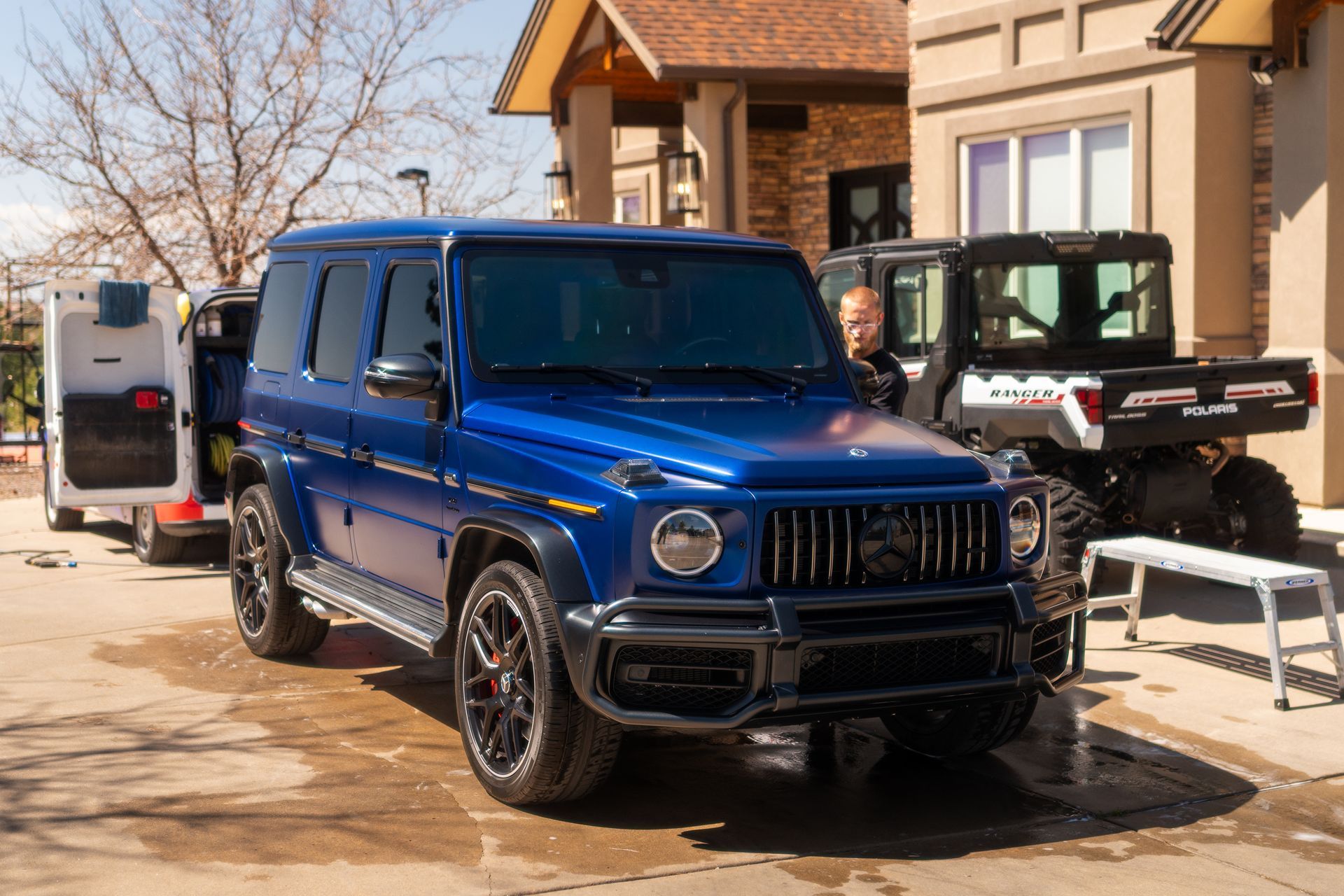 Blue Mercedes SUV in a driveway beside a house, with off-road vehicles and trailer in the background.