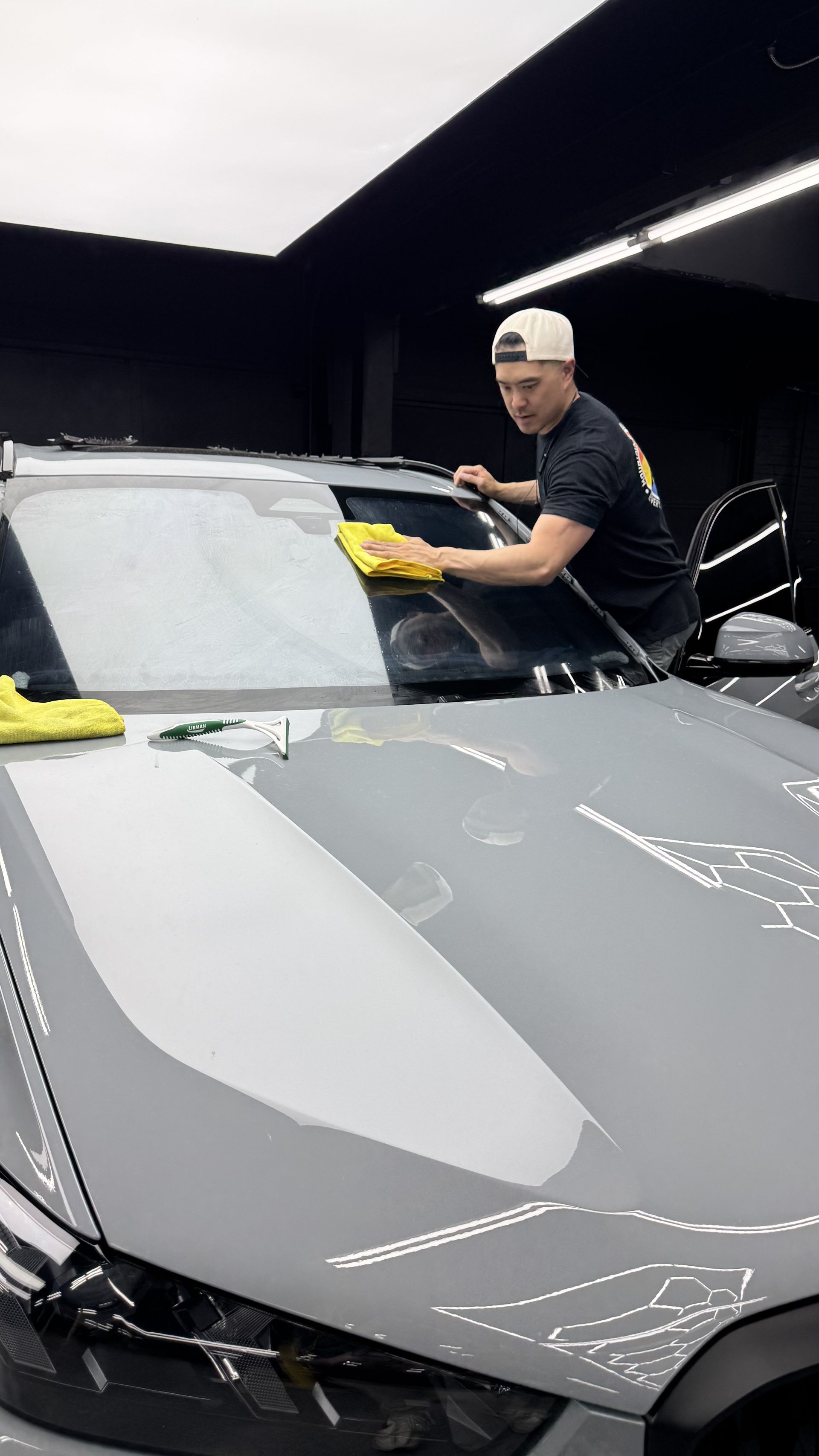 Person polishing a gray sports car in a garage with a yellow cloth