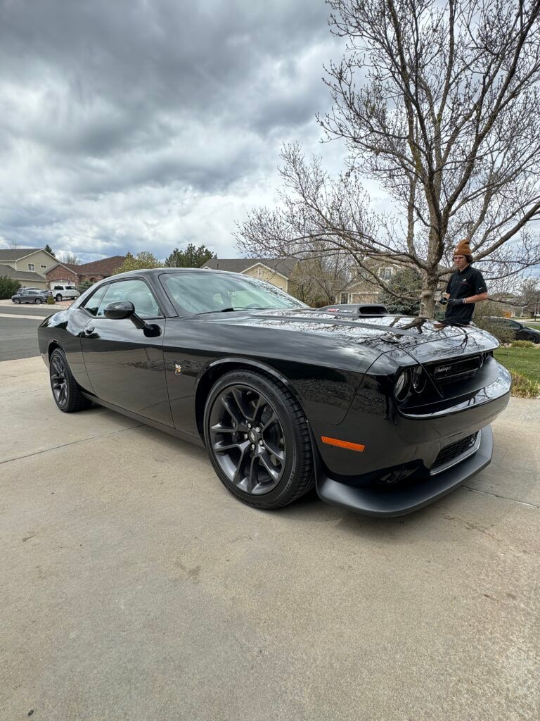 A man is standing next to a black dodge challenger parked on the side of the road.