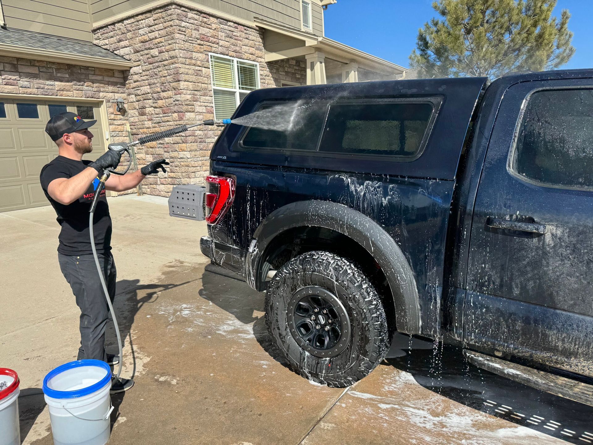 Man washing a black pickup truck with a pressure washer in a driveway on a sunny day.