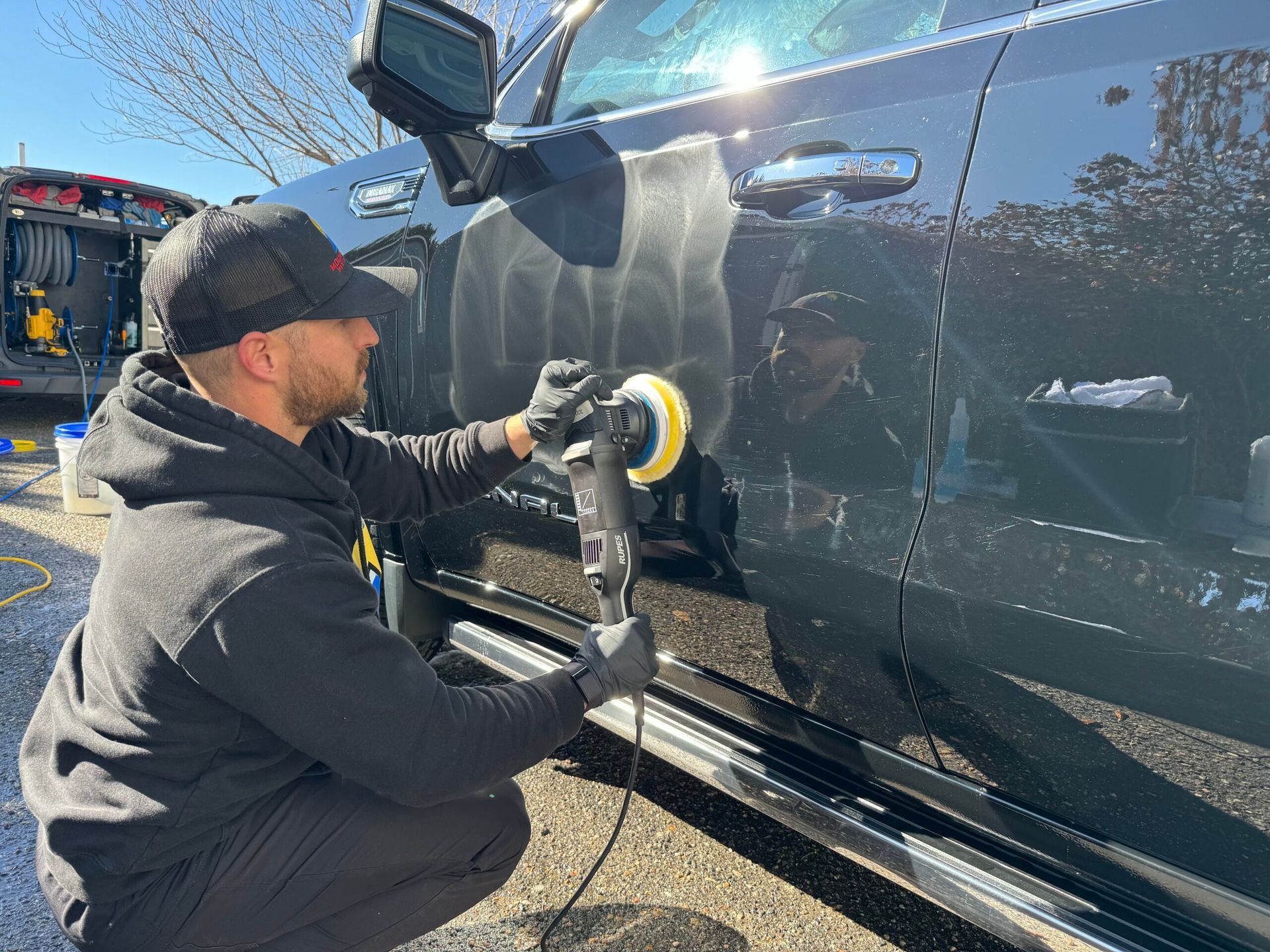 Person polishing a dark-colored vehicle with a power buffer outdoors on a sunny day.