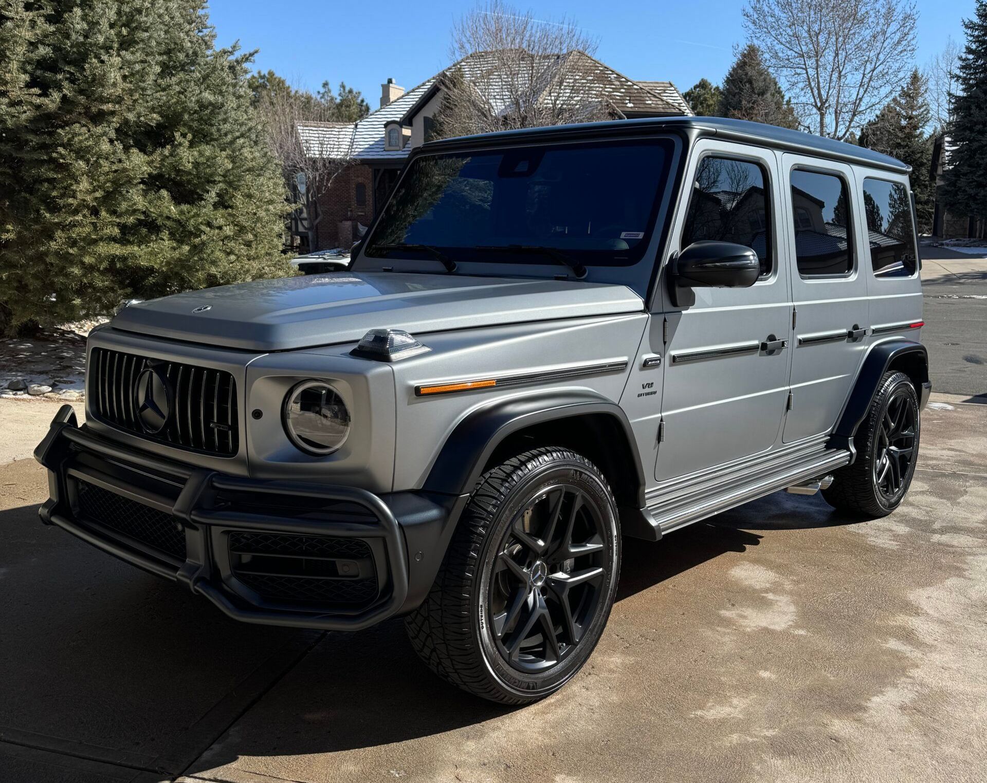 Silver Mercedes-Benz G-Class SUV parked on a driveway with black wheels, under a sunny sky.