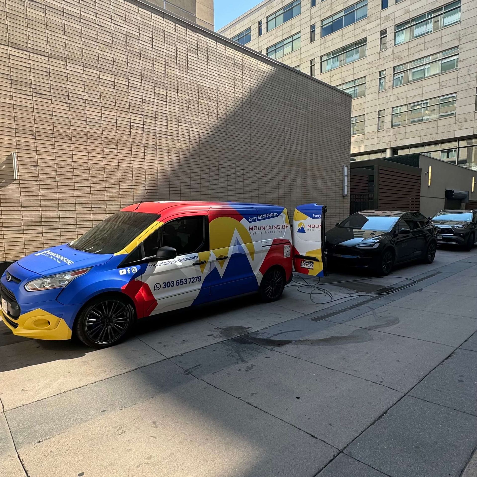 Colorful van with Colorado flag design parked next to black cars in an alley.