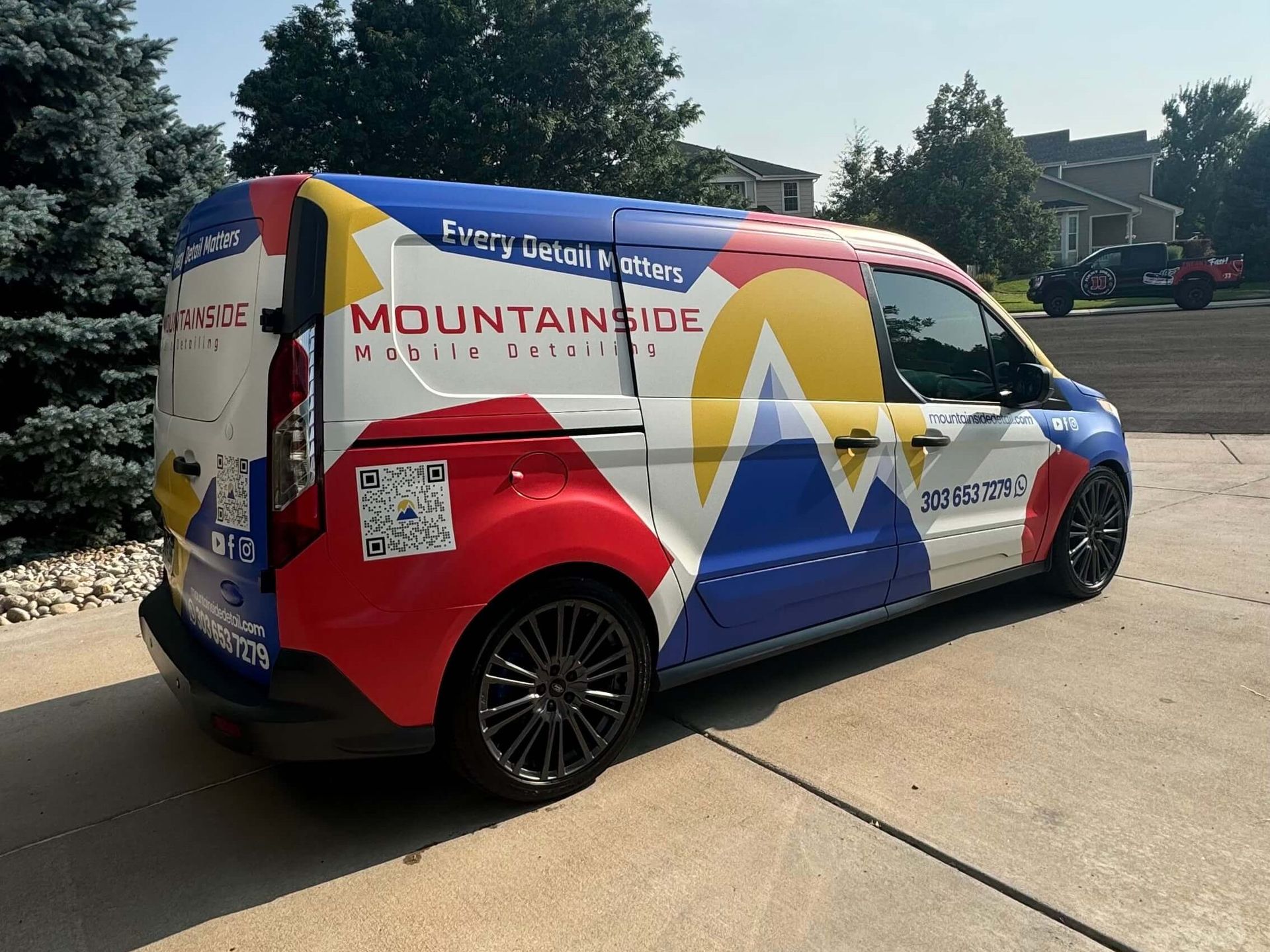 A white van with a colorful mountainside logo and contact info parked outside on a sunny day.