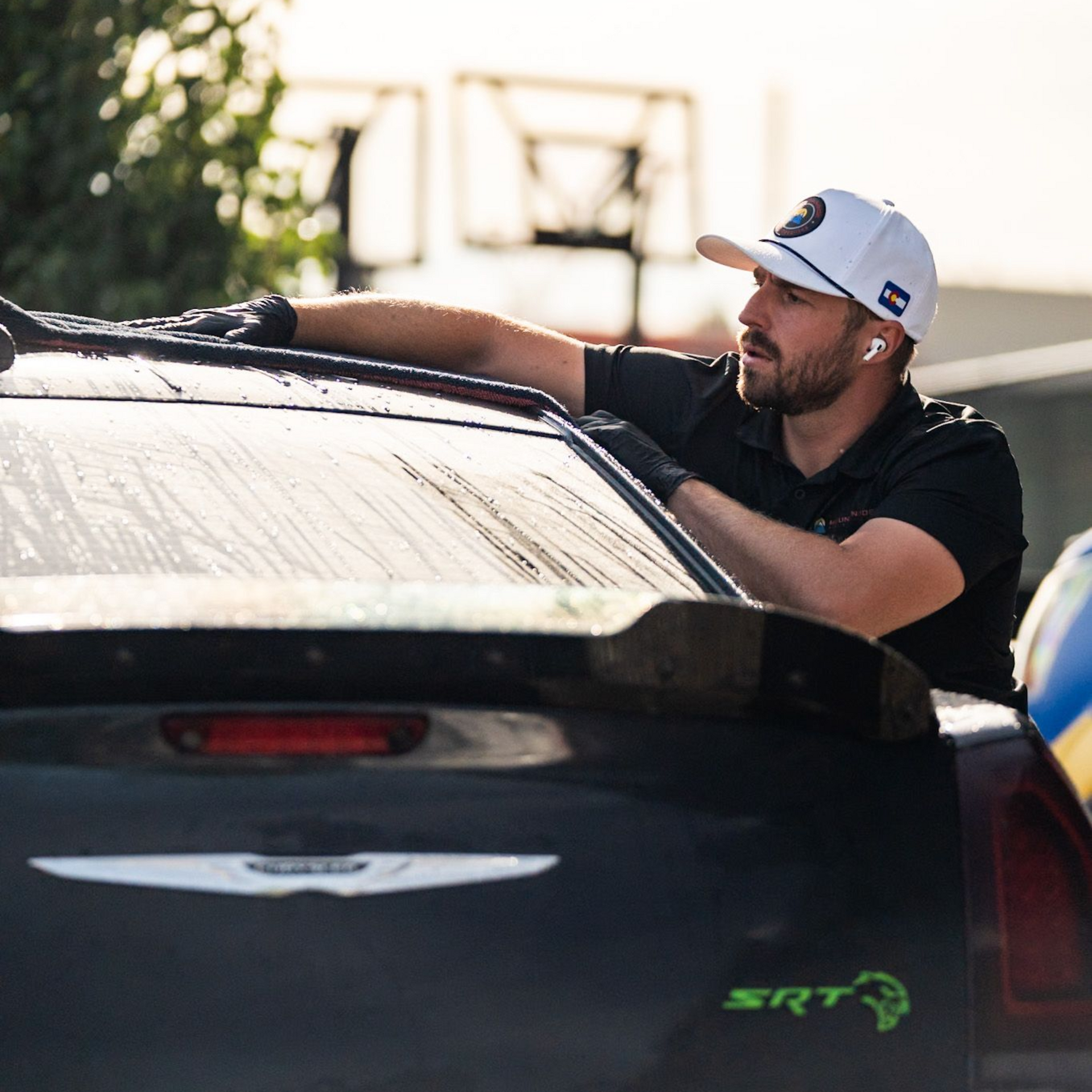 Man leaning on the rear of a black Dodge SRT car, wearing a white cap in bright sunlight