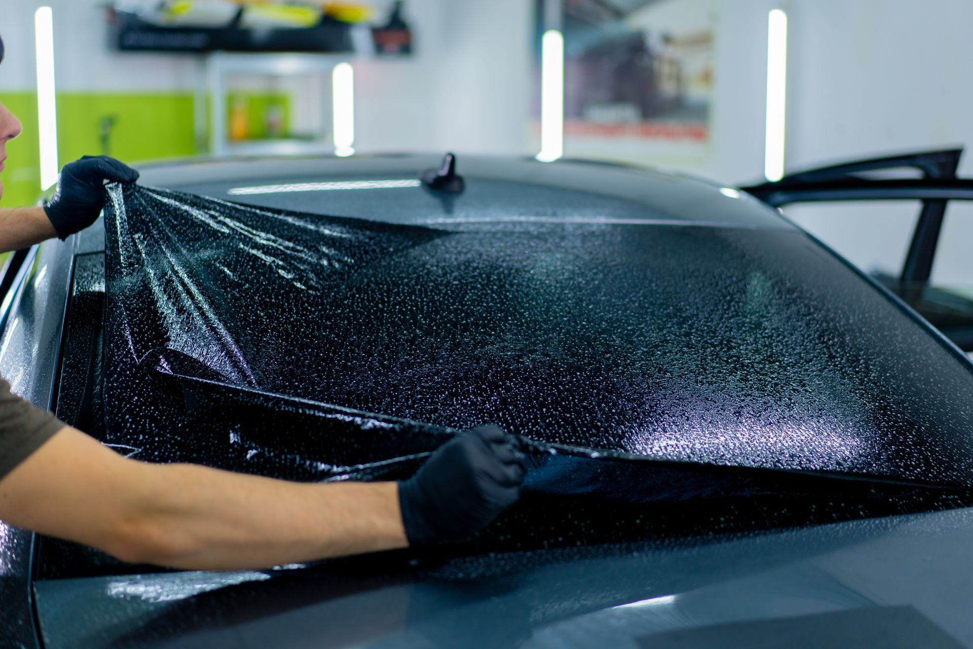 Person applying tint film to a car's rear window in a workshop, using gloved hands.