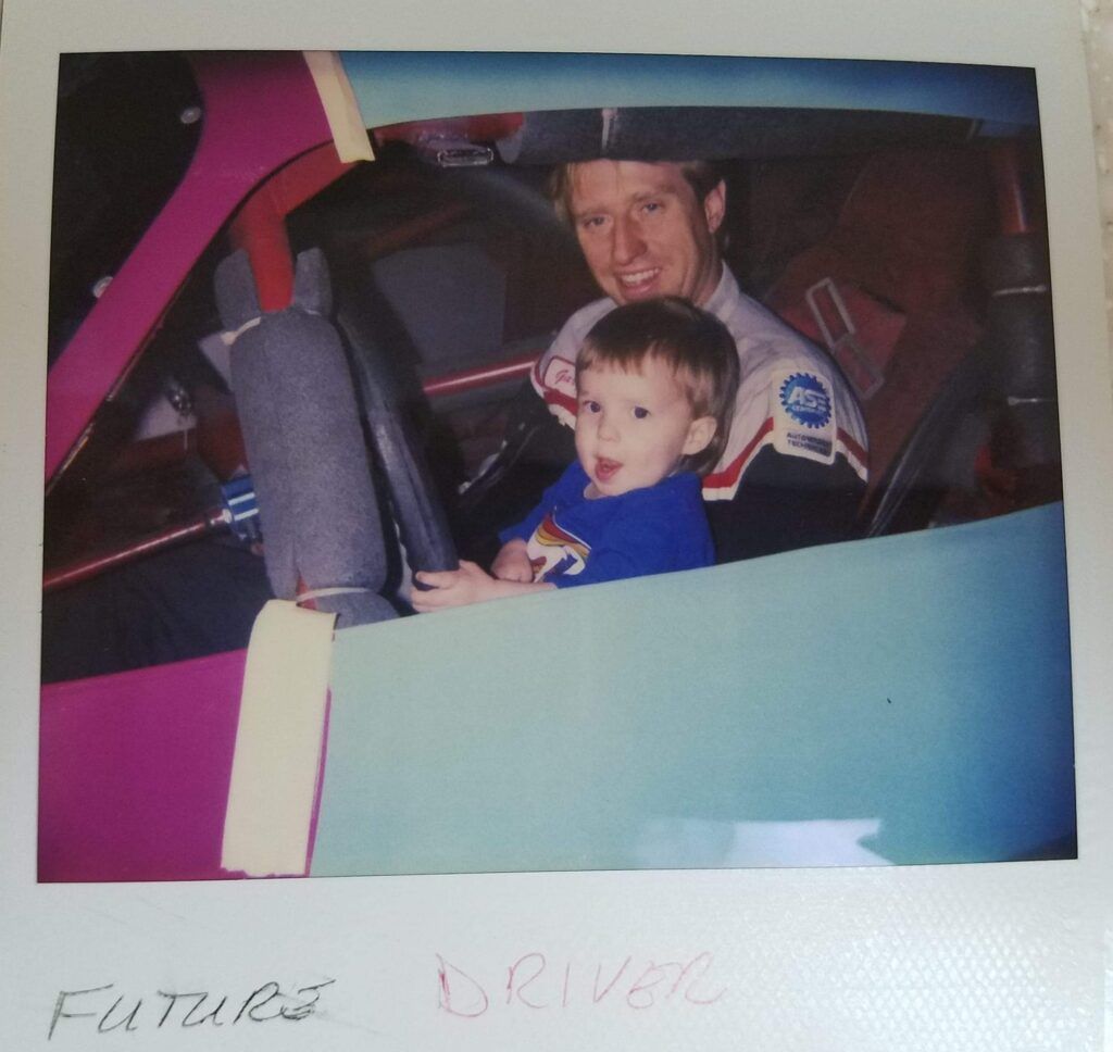 Man and young child in race car. Child sits in the driver's seat, smiling, with the man.