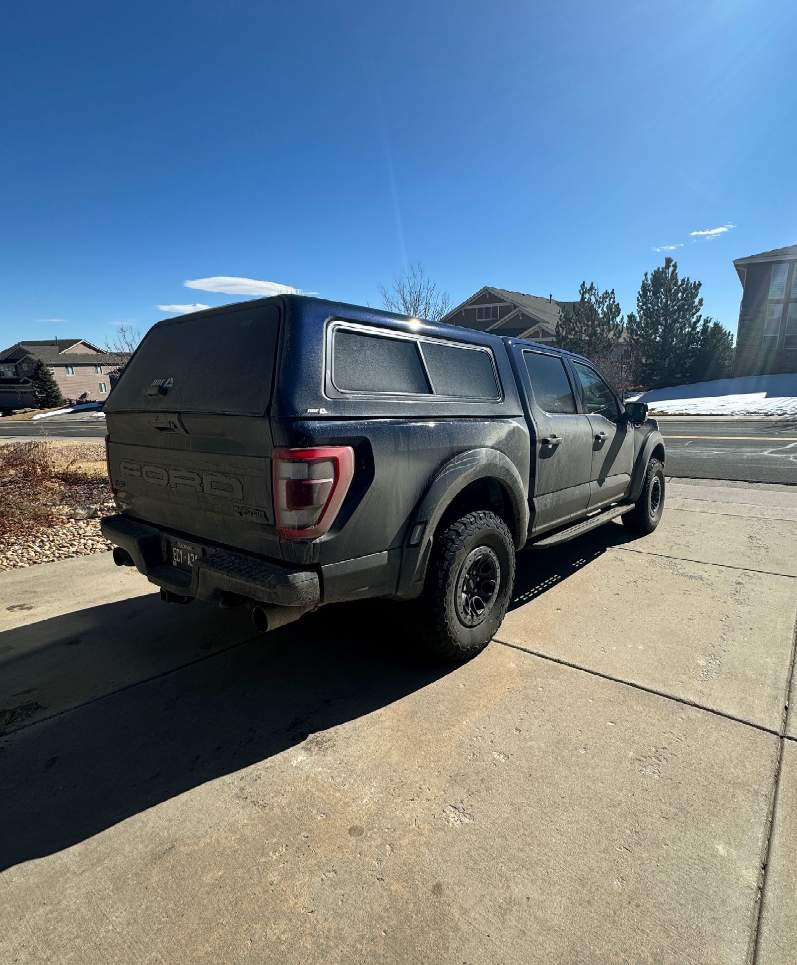 Dark pickup truck with camper shell parked on a driveway in a sunny outdoor setting.