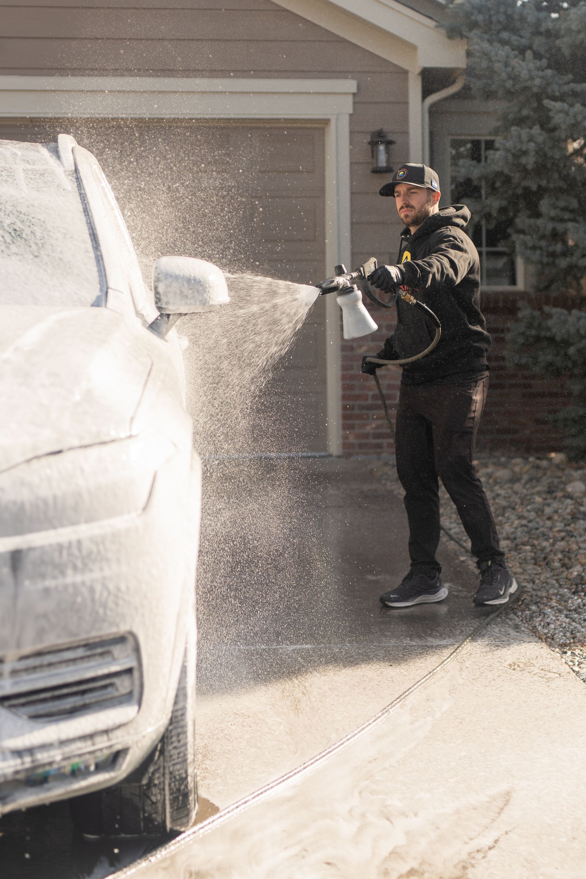 Man sprays car with foam wash in a driveway near a garage.