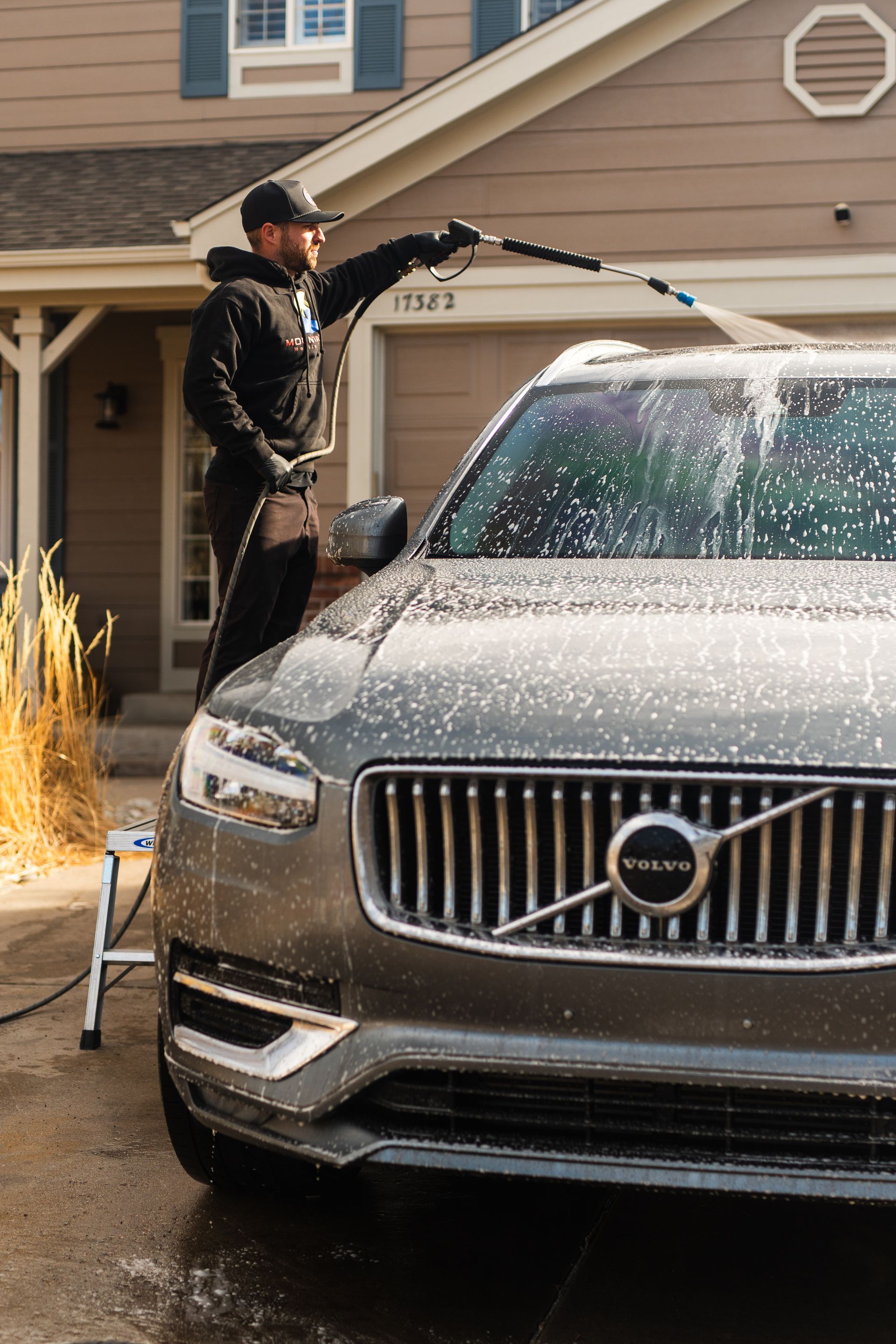 Person washing a gray Volvo SUV with soap and water in a driveway.