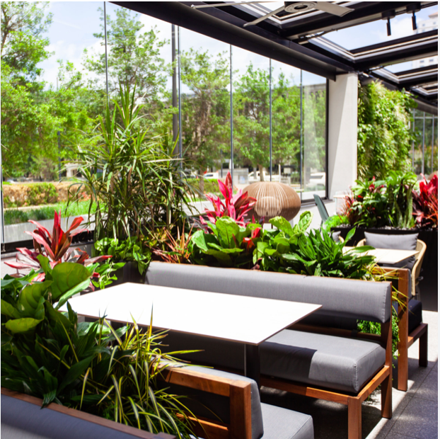 Outdoor dining area with lush greenery, gray and white seating, and glass walls.