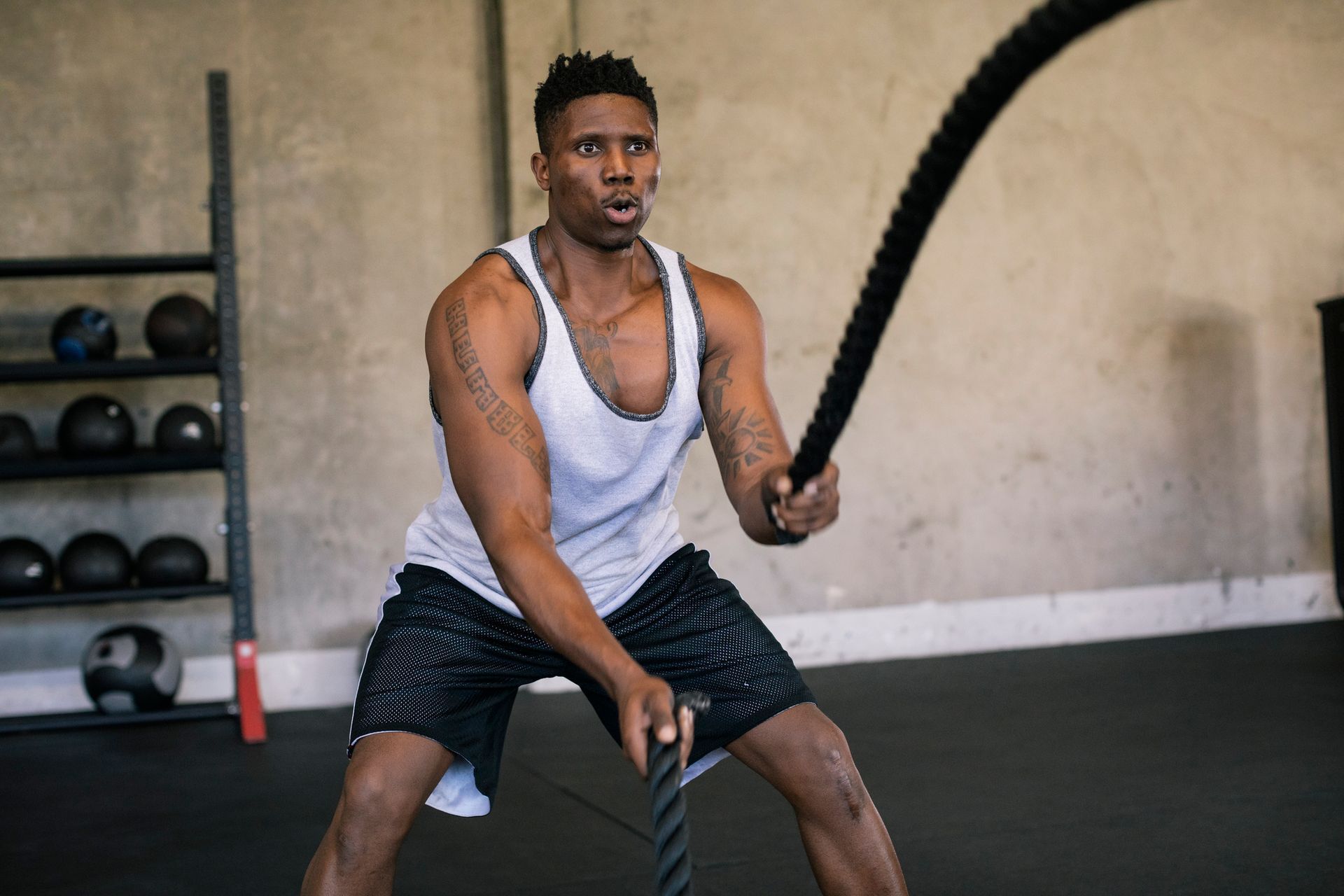 Man exercising with battle ropes in a gym, wearing a white tank top and black shorts.