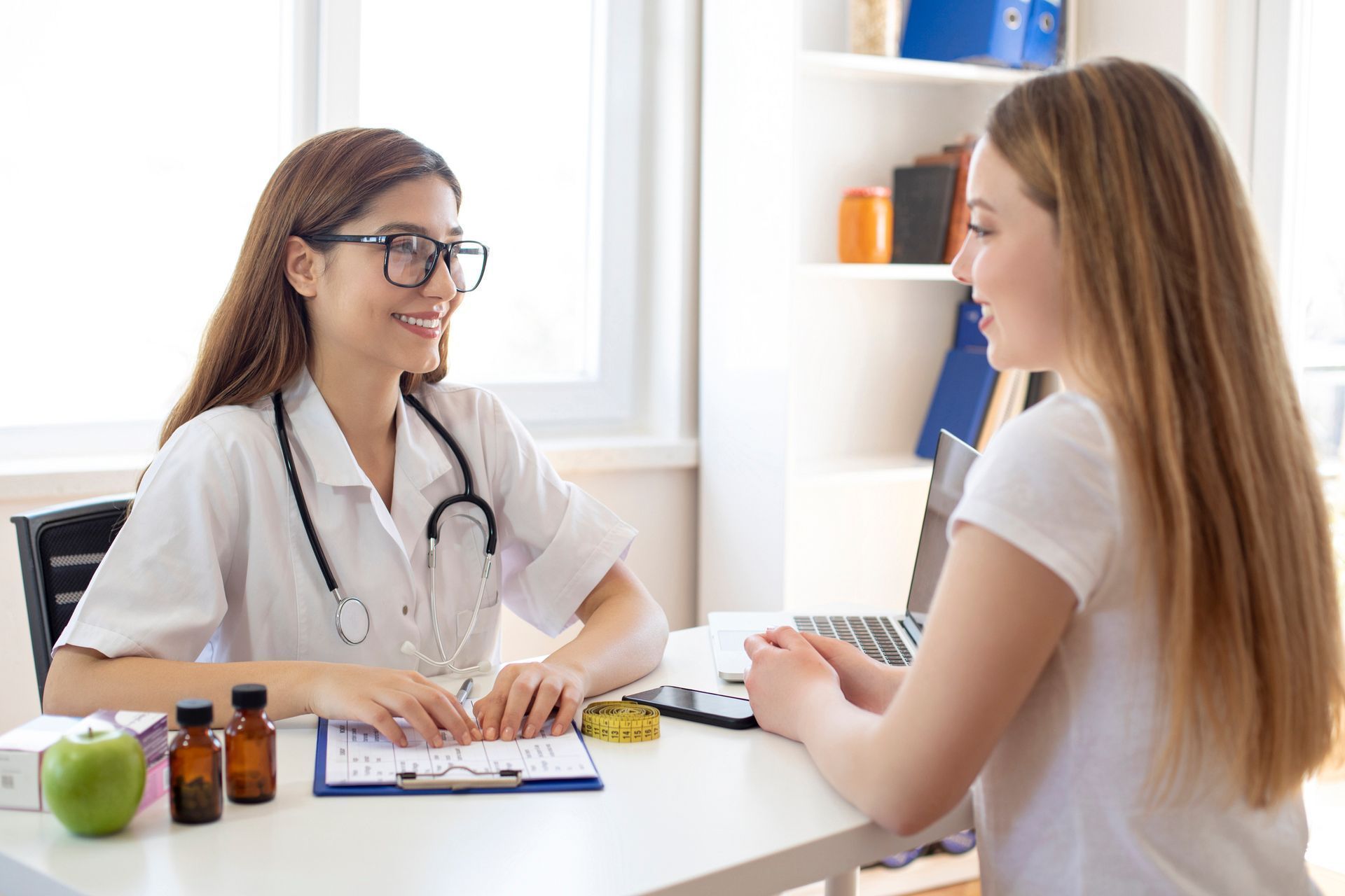 Doctor consulting with a patient at a desk in a bright clinic, with a laptop and medicines nearby.