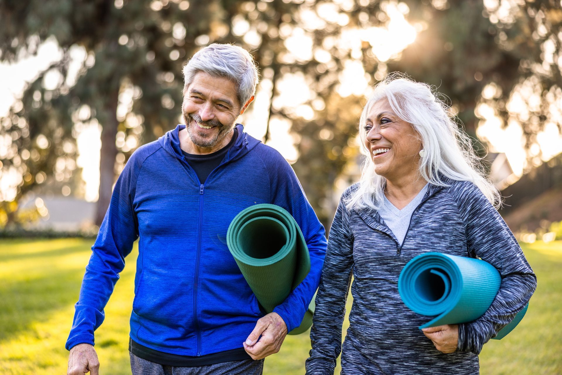 Two smiling people walking outdoors with rolled yoga mats in a sunny park