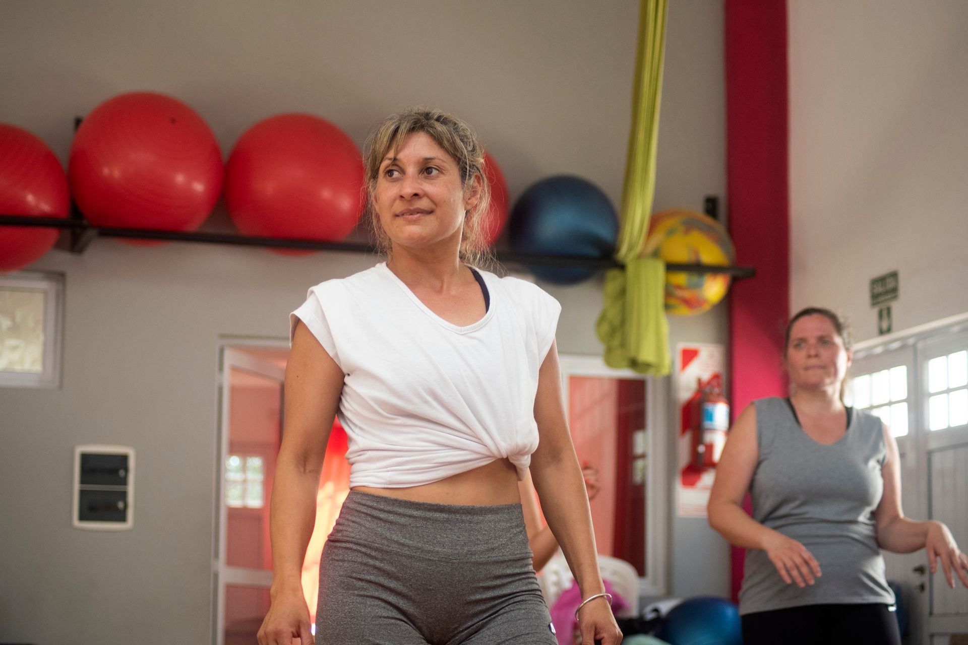 Two women in a gym, one in a white top and one in gray, standing near exercise balls.