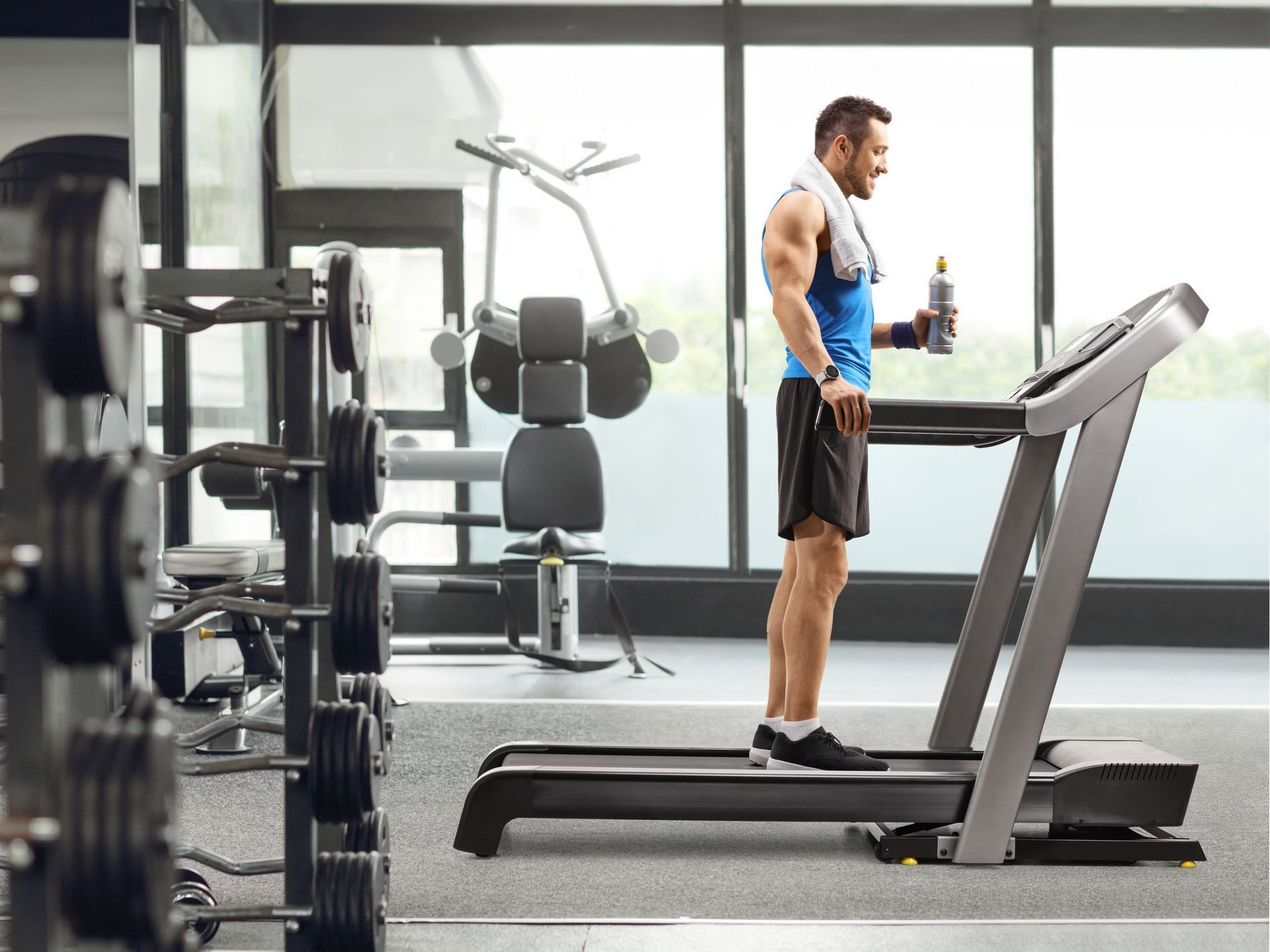 Man standing on a treadmill in a bright gym beside strength machines and dumbbells