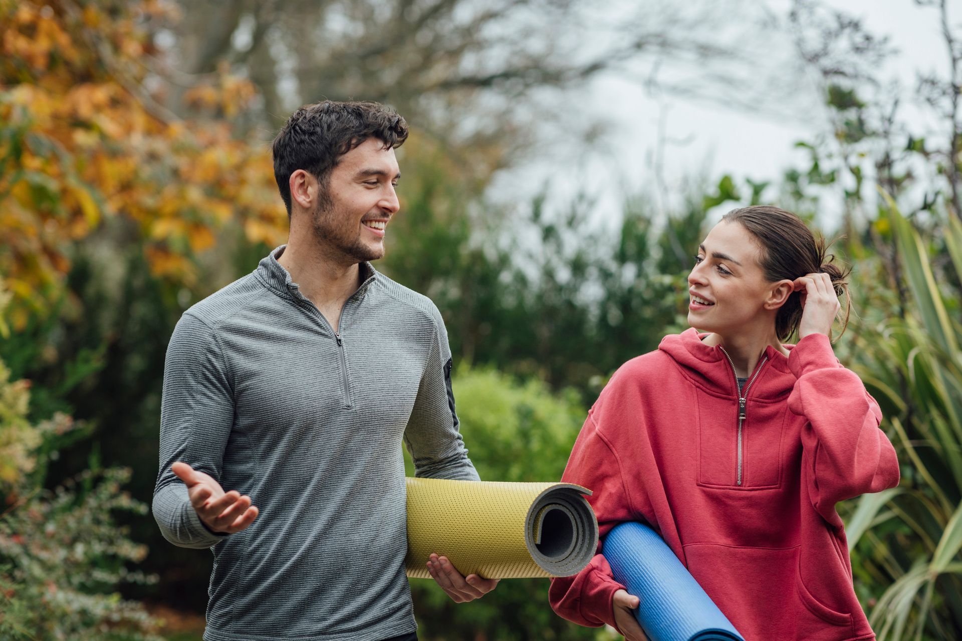 Two people in outdoor gear walk and talk, carrying rolled-up mats in a green park.