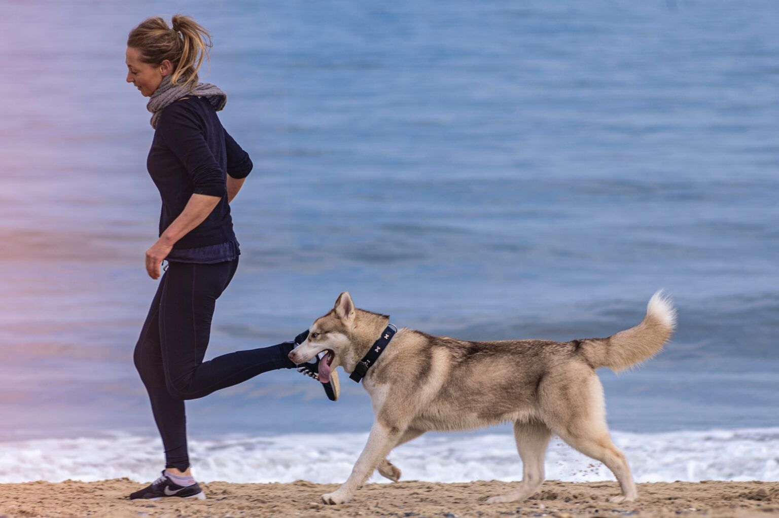 Woman running on a beach beside a dog on a leash, with the ocean in the background