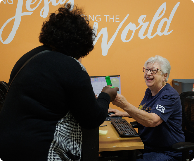 Two people at a desk exchanging a green card, with a smiling woman seated before an orange wall sign.