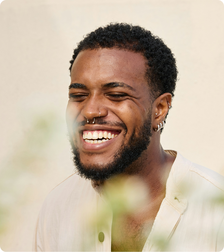 Smiling man with short curly hair and beard, wearing a light shirt outdoors, softly blurred background